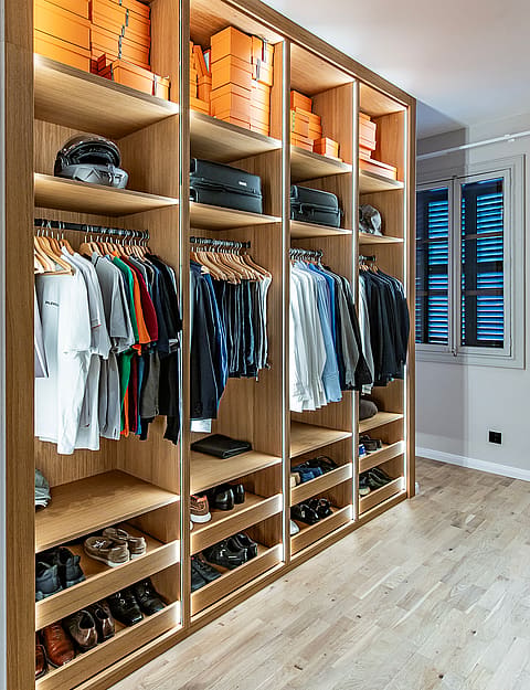 A walk-in dressing room featuring light oak cabinetry with illuminated shelving, hanging rails, and shoe racks, leading to an adjacent bedroom.