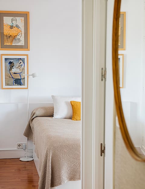 A close-up view of a bedroom interior with a bed featuring textured bedding, two framed artworks on the white wall, and a large oval mirror in the foreground.
