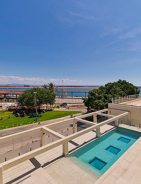 Vista en ángulo de una terraza con piscina rectangular, sobre un parque, la carretera costera y el mar.