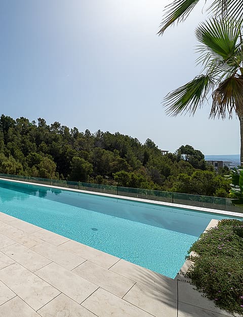 A long rectangular swimming pool with a stone-paved deck, lounge chairs, and a glass balustrade overlooking a tree-lined hill.