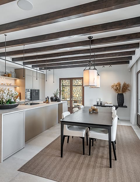 Modern dining area showing dark wood ceiling beams, a central table with white chairs, and an adjacent kitchen island.