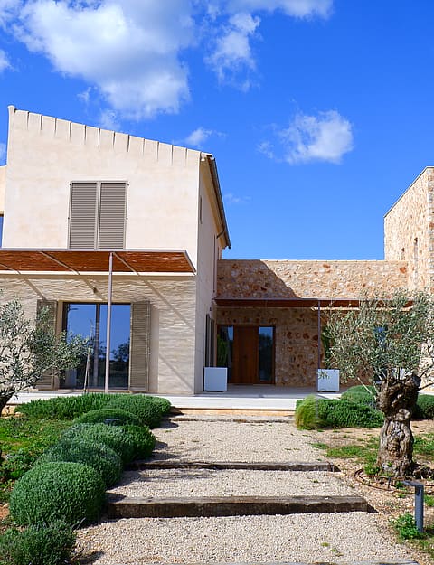 Two-story stone-clad finca with a graveled pathway, mature olive trees, and wooden pergola under a clear blue sky.