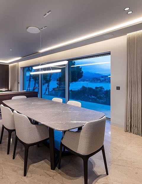 Modern dining room with stone table, upholstered chairs, and floor-to-ceiling glass windows overlooking a coastal landscape.