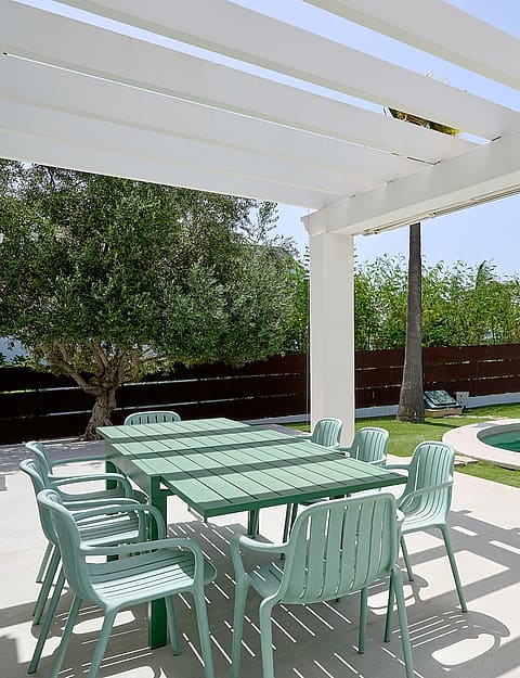 Pérgola blanca sobre mesa de comedor exterior junto a piscina circular con cipreses y setos perfilados