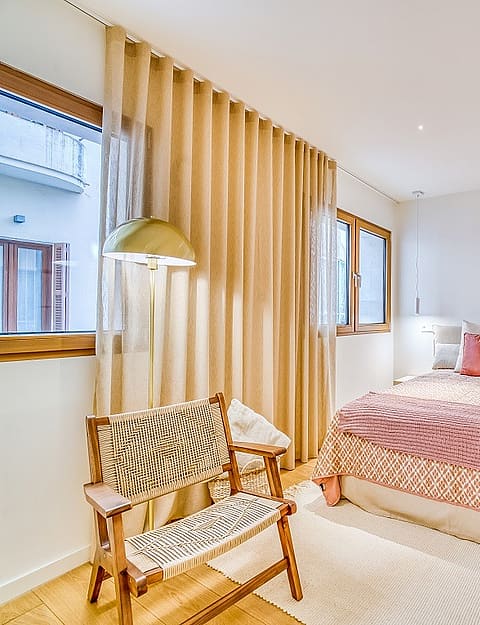 A bright bedroom with light wooden floors, a double bed with coral bedding, and a wooden armchair beside floor-length curtains.