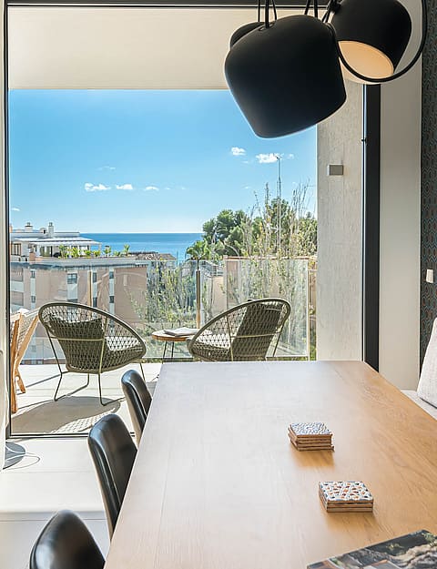 Dining room with wooden table, modern chairs, and black pendant lights, opening to a terrace with wicker armchairs and sea views.