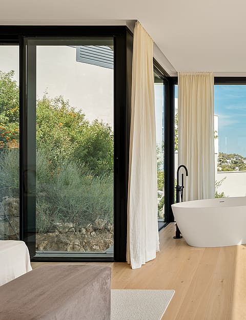 A modern master bathroom features a white freestanding oval bathtub, light oak hardwood floors, and floor-to-ceiling black-framed windows overlooking a lush garden.