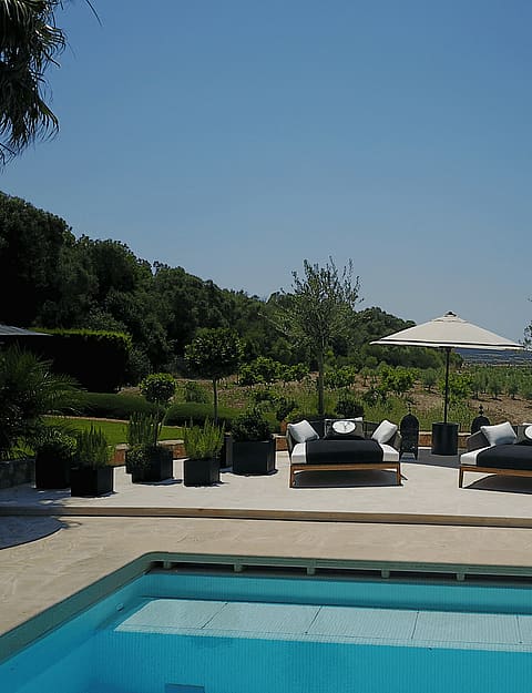 A swimming pool deck with two black and white lounge chairs and sun umbrellas, framed by olive trees and open landscape.