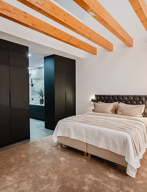 A modern bedroom with a large bed against a white wall, dark floor-to-ceiling wardrobes, and visible natural wood ceiling beams.