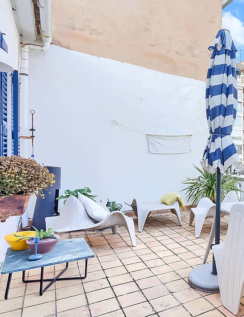 A sunny outdoor terrace with a dining table, chairs, and a blue-striped parasol, featuring city views of neighboring buildings.
