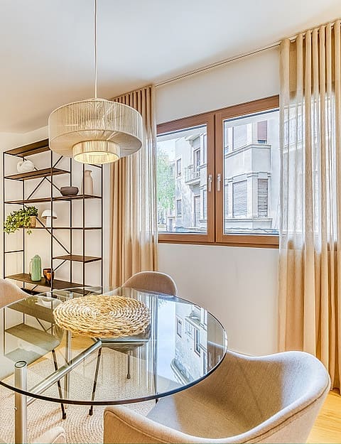 A glass dining table with four curved chairs next to a metal shelving unit in an apartment, with linen curtains framing city windows.