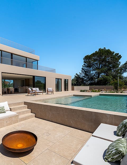 A modern villa exterior featuring a sunken stone lounge with built-in seating, a circular fire pit, and an adjacent swimming pool under a clear blue sky.