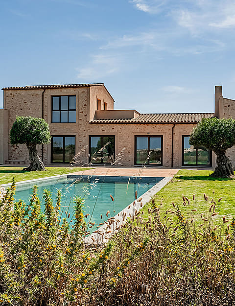 Exterior view of a traditional stone finca with a swimming pool, manicured lawn, and native trees under a clear blue sky.