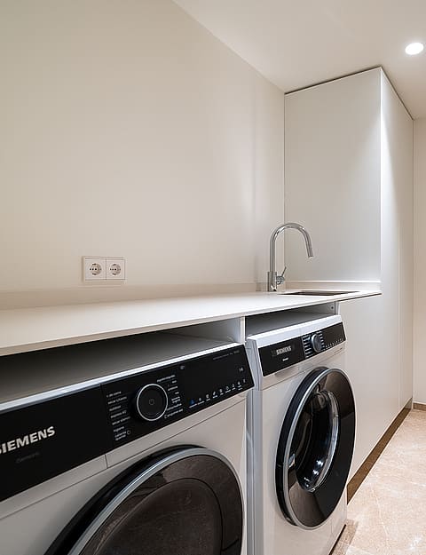 A white minimalist laundry room featuring a stacked washer and dryer, white countertops, a small steel sink, and light flooring.