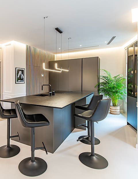 Modern kitchen featuring a dark grey central island, black swivel stools, recessed ceiling lighting, and panelled walls.