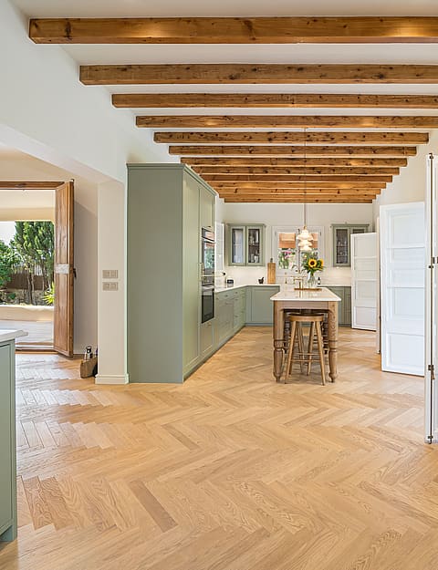 Open-plan kitchen featuring sage green cabinets, light oak herringbone floors, and exposed ceiling beams.