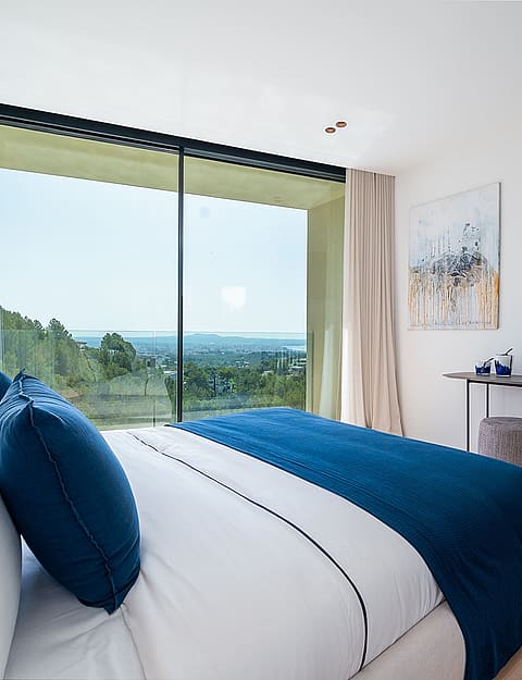 Modern bedroom with a large floor-to-ceiling window overlooking a coastal landscape, featuring a white bed with blue pillows.
