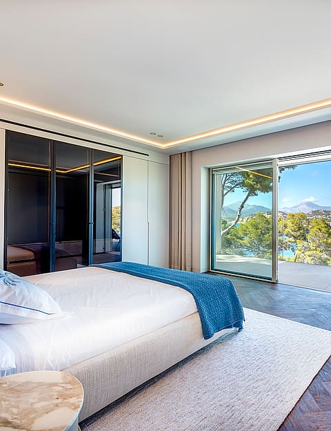 Modern bedroom with dark herringbone wood floors, a large white bed, and floor-to-ceiling glass sliding doors to a terrace.