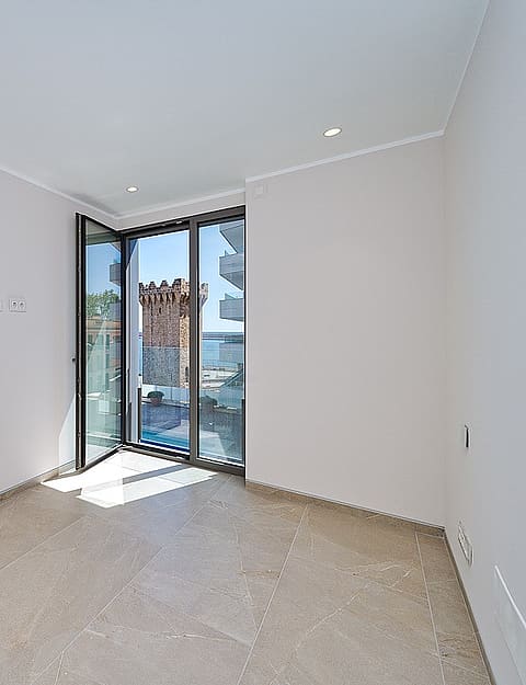 Bright unfurnished bedroom with pivot glass door opening to balcony, Torre de Paraires and harbour visible beyond