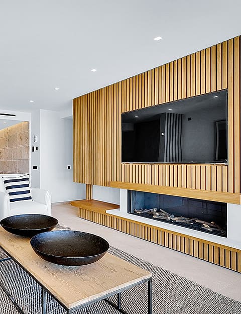 Interior view of a living room featuring a vertical oak slatted wall with an integrated fireplace and TV, white sofas, and coffee table.