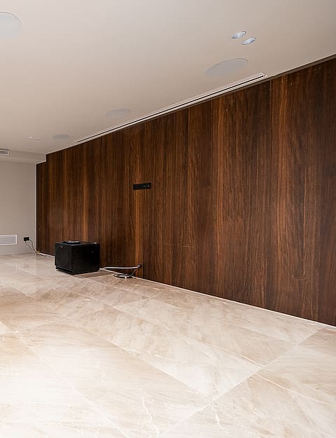 A minimalist home cinema room featuring polished beige stone floors and a dark wood-paneled accent wall, with a ceiling-mounted projector.