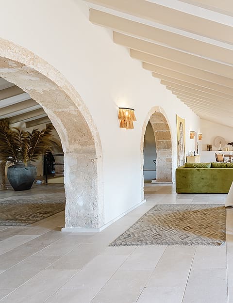A spacious bedroom with a king-sized bed, tufted headboard, stone flooring, and arched doorways leading to an adjoining area.