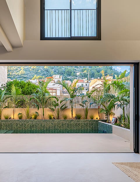 Interior view through open sliding doors to a terrace with a green tiled plunge pool and lush perimeter planting.