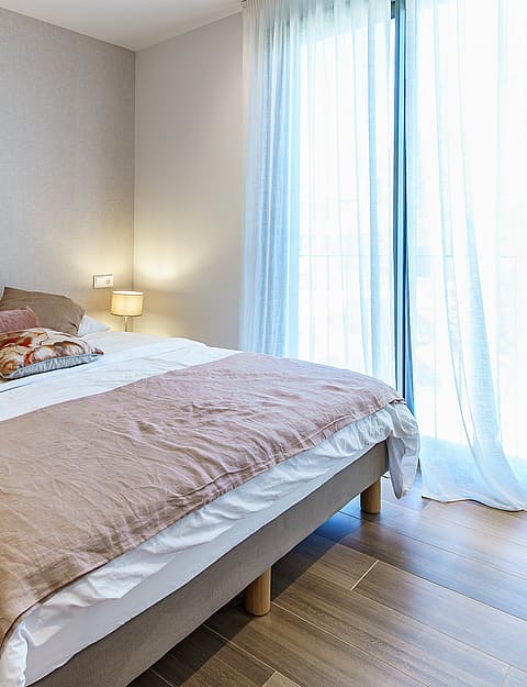 A bright bedroom with a king-sized bed, beige textile headboard, mirrored side tables, and sheer white curtains.
