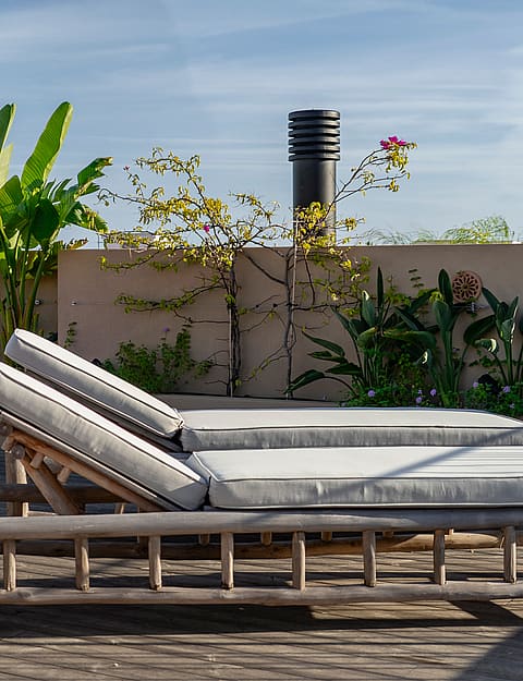 A rooftop terrace in Genova features two wooden sun loungers with grey cushions placed against a stone planter wall with plants.