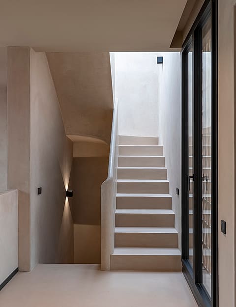 A neutral-toned minimalist staircase with smooth plaster walls, recessed wall lighting, and a dark metal door frame.