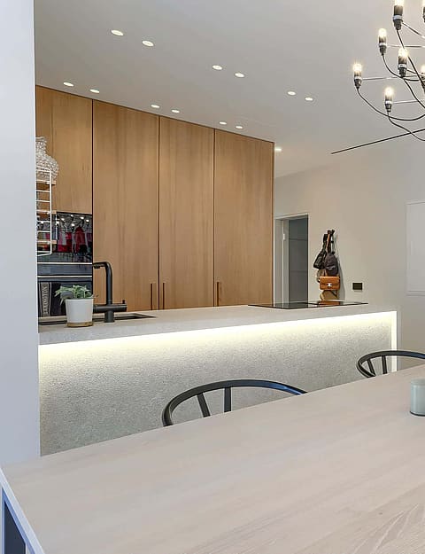 Dining room with light oak table and black chairs beside an open kitchen with illuminated stone island and modern chandelier.