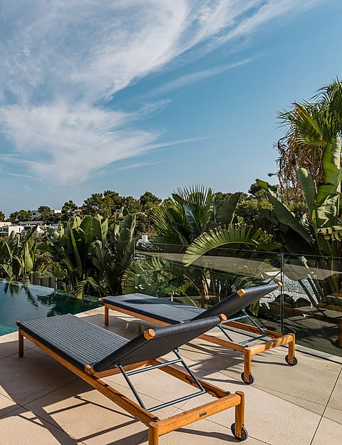 Two dark wicker sun loungers with wooden frames on a stone terrace overlooking an infinity pool and Mediterranean foliage.