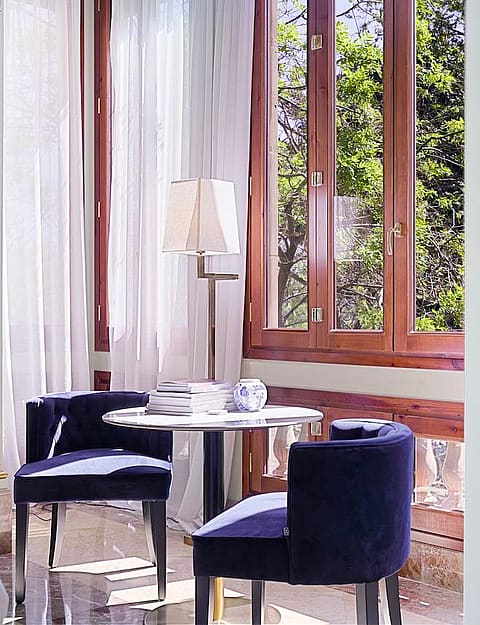 A small circular table with two blue upholstered chairs placed beside tall wood-framed windows with white curtains.