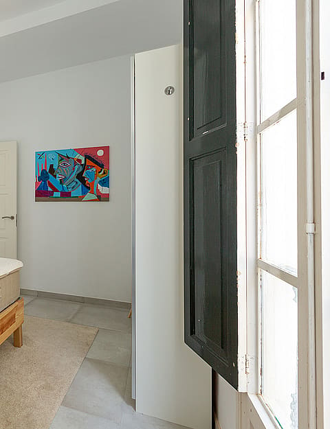 A bright bedroom with white walls, a single wooden bed frame, and an open window with dark shutters.
