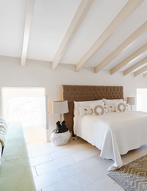 Spacious bedroom with a tufted headboard, white linens, and light wood rafters in a bright, minimalist attic space.