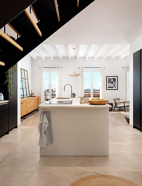 An open-plan kitchen in a Santa Catalina apartment featuring black cabinetry, a white central island, and a dark metal staircase.