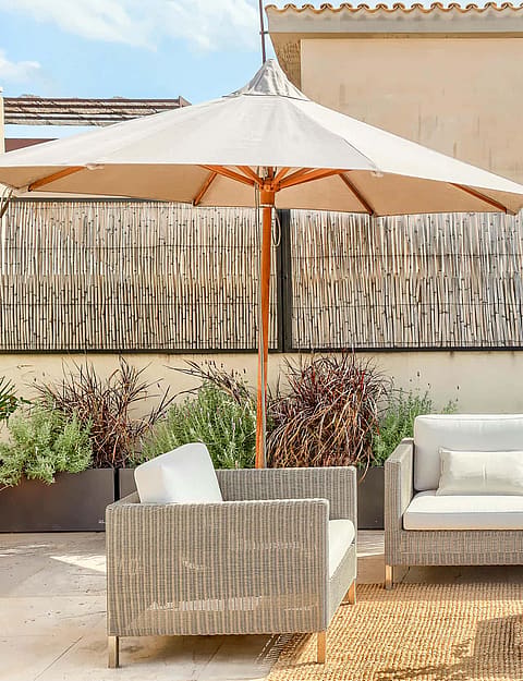 A rooftop terrace featuring a modular wicker sofa set with white cushions, a matching coffee table, and a large beige parasol, set against a backdrop of Old Town buildings.