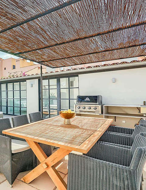 A spacious outdoor terrace with a wooden dining table, wicker chairs, and a shaded lounge area against a city backdrop.