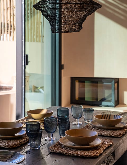 A close-up view of a dining table set with rustic ceramic bowls, blue glassware, and woven placemats, featuring a textured pendant lamp above.