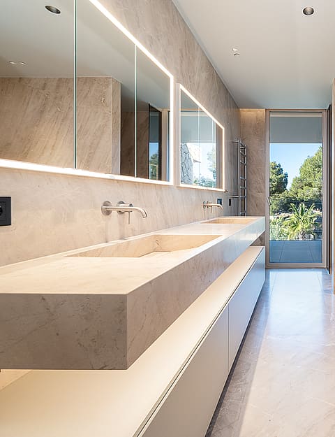 Modern bathroom featuring a long, wall-mounted stone double vanity, integrated LED lighting, and a glass-paneled garden view.