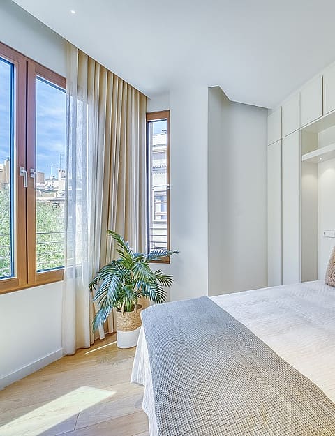 A bright bedroom with a double bed, integrated ceiling-height white wardrobes, and windows showing an urban view.