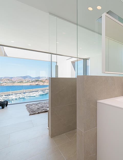 A modern bathroom featuring a white floating vanity, floor-to-ceiling glass walls, and an expansive view of the sea and harbor.