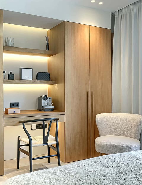 A modern bedroom featuring a floor-to-ceiling light oak wardrobe unit with a built-in desk, shelving, and a black wishbone chair.