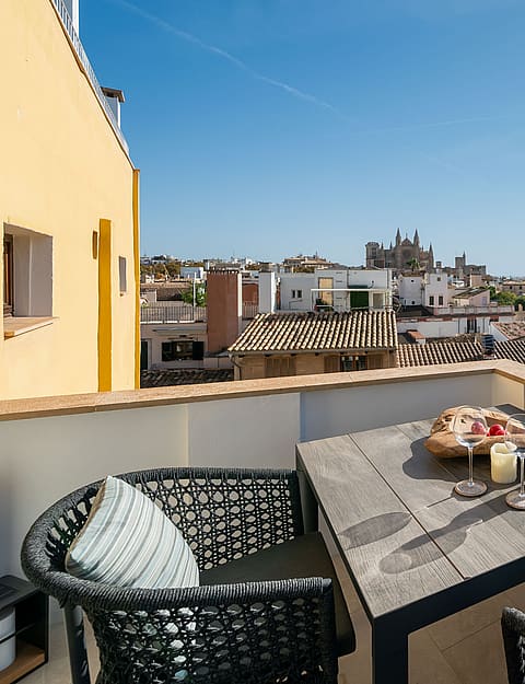 Rooftop terrace with wooden dining table and woven chairs overlooking historic Palma rooftops and the cathedral.