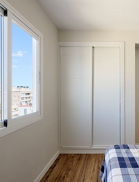 A minimalist bedroom featuring light walls, a wooden floor, and built-in sliding wardrobes, with a window view of city buildings.