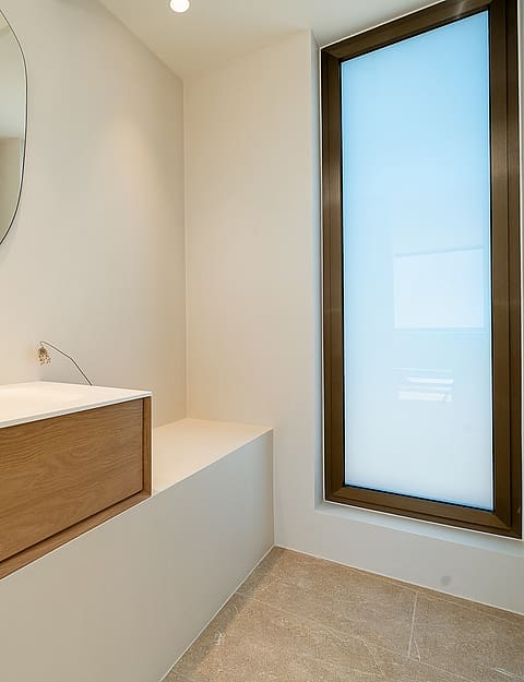 Modern bathroom with a wall-mounted oak vanity, circular mirror, and tall frosted window allowing diffused natural light.