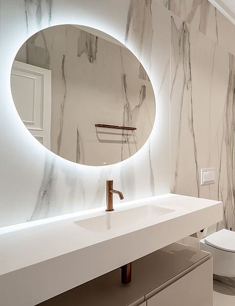 A modern bathroom featuring floor-to-ceiling white marble with grey veining, a wall-mounted vanity, and a backlit circular mirror.