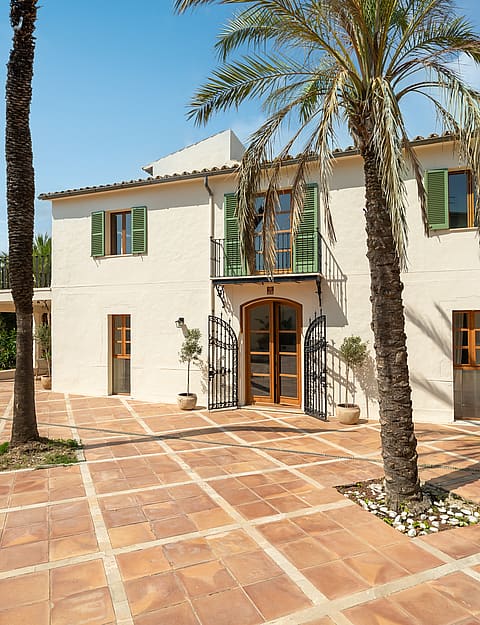 Cream stucco villa with green shutters and an arched wooden door opening onto a geometric terracotta tile courtyard.