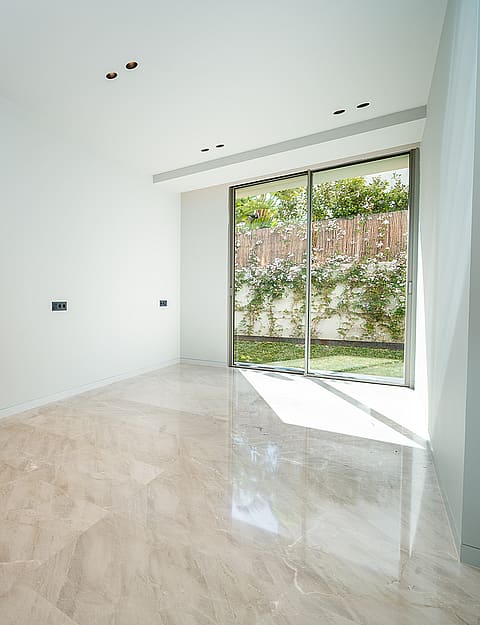 Empty rectangular room with polished marble floors, plain white walls, and a large sliding glass door opening to a garden.
