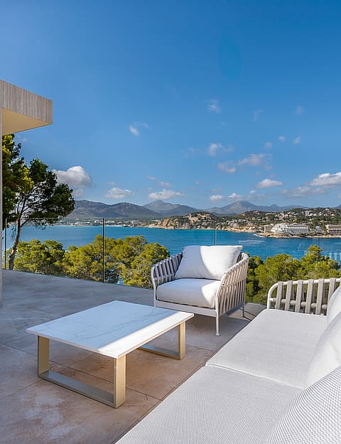 Outdoor terrace with white lounge furniture, glass railings, and a wide view of the Mediterranean bay and coastal mountains.